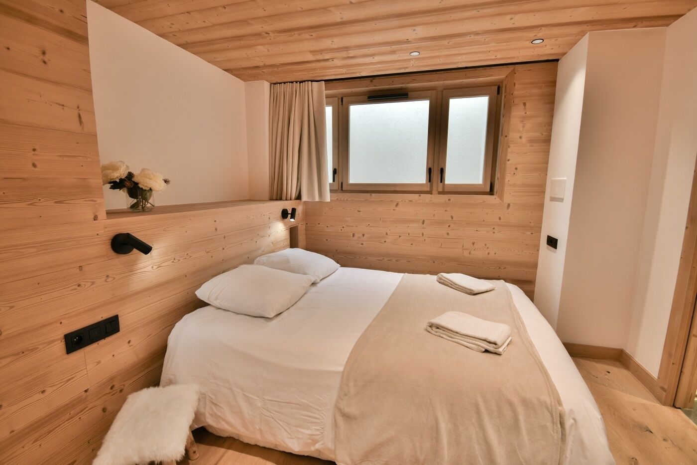 A bedroom with a wooden headboard, a bed with white bedding, a window with curtains, and a small bedside table with a lamp.