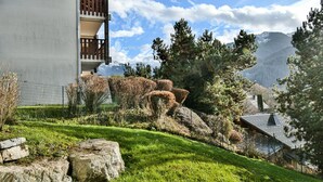 Una casa en una ladera con balcón de madera, rodeada de un jardín cuidado y vistas a una cadena montañosa.