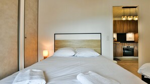 A hotel room with a bed, a nightstand with a lamp, a wooden headboard, and a view of the kitchen area. - Le Sève - B103 - One Bedroom Apartment, Sleeps 5 (Samoëns)