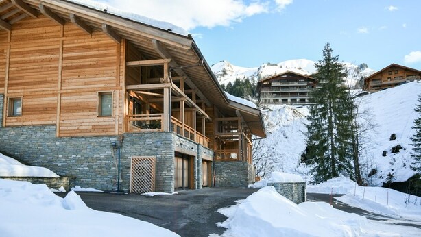 Un chalet en bois aux murs de pierre, doté d’un balcon et offrant une vue sur des montagnes enneigées.