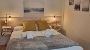 Un lit bien fait, recouvert d’une couverture grise, avec deux oreillers de couleur moutarde et une serviette blanche pliée posée sur le lit. On y trouve deux tables de chevet avec des lampes, une œuvre d’art accrochée au mur et un tableau encadré au-dessus du lit.