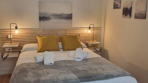 A neatly made bed with a grey blanket, two mustard-colored pillows, and a white towel folded on the bed. There are two bedside tables with lamps, a wall-mounted artwork, and a framed picture above the bed. - 135A1 Charming Apartment in Gijón by R2R (Gijón)