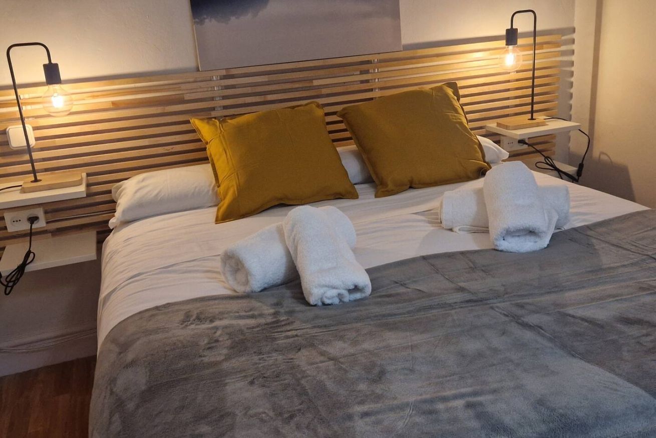 A neatly made bed with two pillows, a grey blanket, and two rolled towels.