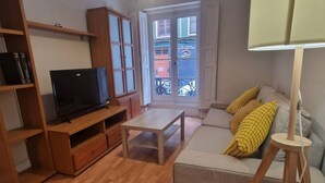 A living room with a television, wooden furniture, a sofa with cushions, and a window with a view of buildings. - 135A1 Charming Apartment in Gijón by R2R (Gijón)