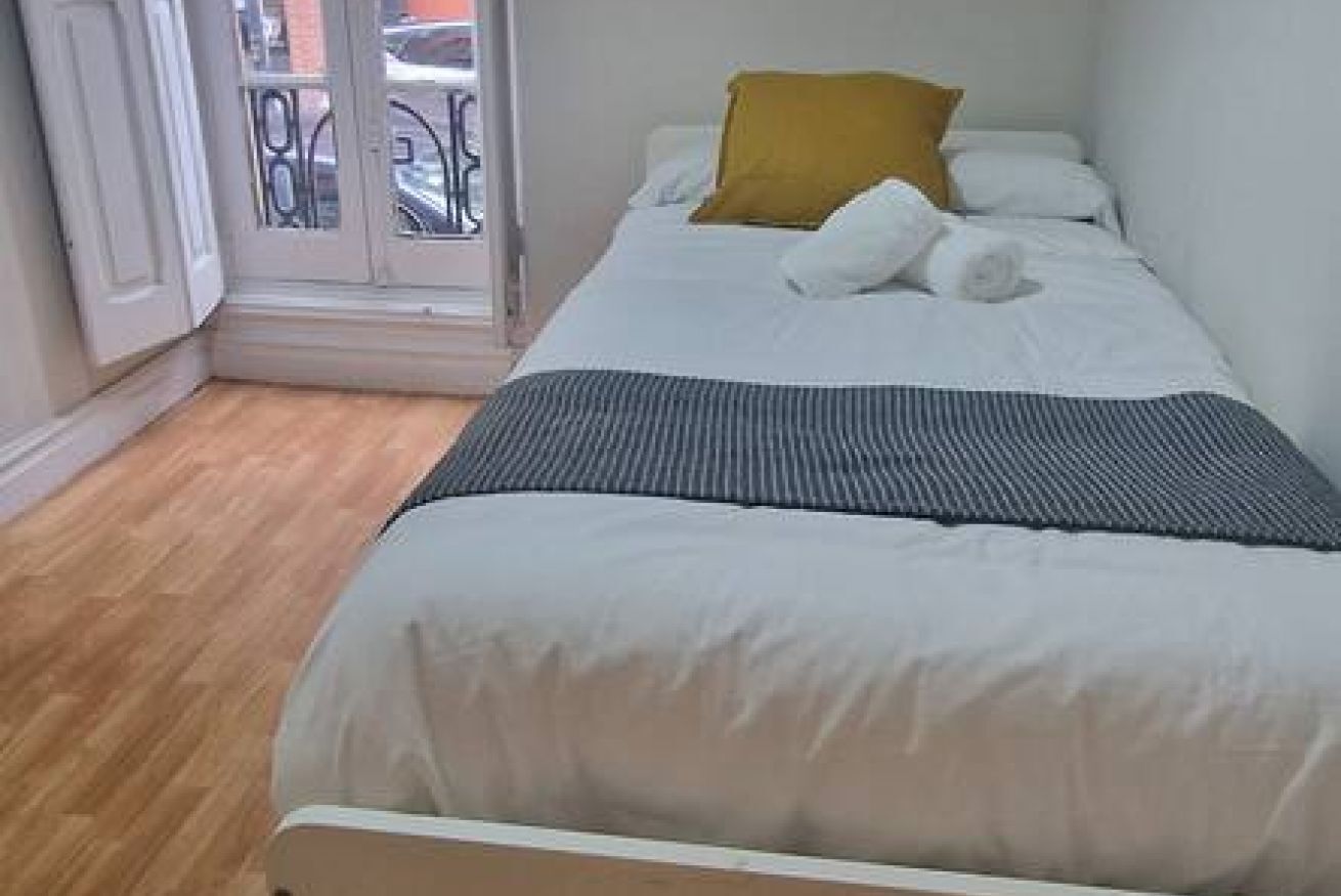 A single bed with white bedding and a mustard-colored pillow, a wooden floor, and a door leading to an outdoor area.