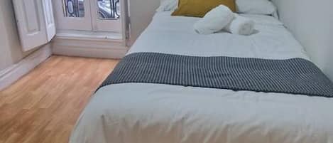 Ein Einzelbett mit weißer Bettwäsche und einem senfgelben Kopfkissen, Holzboden und eine Tür, die zu einem Außenbereich führt.