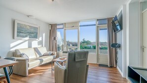 Ένα σύγχρονο σαλόνι με έναν καναπέ, μια πολυθρόνα και μια τηλεόραση, μεγάλα παράθυρα με κουρτίνες και έναν πίνακα στον τοίχο.