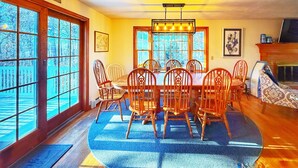 A dining area with a wooden table and chairs, large windows, and a fireplace. - Stevie, The Cape Farmhouse (Chatham)