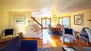 A spacious living room with a wooden staircase, a dining area with a wooden table and chairs, and a bookshelf filled with books. - Stevie, The Cape Farmhouse (Chatham)