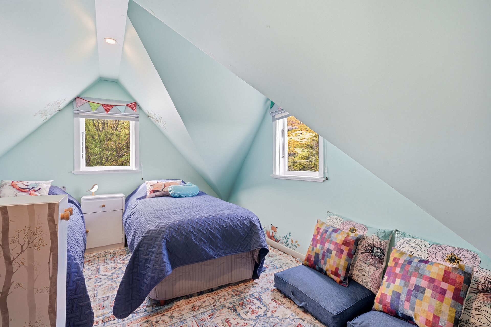 Un dormitorio con techo inclinado, dos ventanas, una cama con colcha azul, una mesita de noche, un aparador y una alfombra colorida.