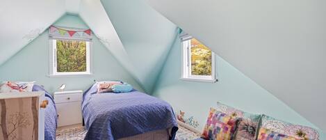 Un dormitorio con techo inclinado, dos ventanas, una cama con colcha azul, una mesita de noche, un aparador y una alfombra colorida.