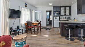 Une cuisine moderne avec des meubles de couleur sombre, un four à micro-ondes, un réfrigérateur et un grille-pain. On y trouve également un coin repas avec une table et des chaises en bois, ainsi qu’un téléviseur fixé au mur.