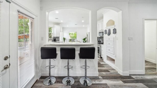 A modern kitchen with white cabinetry, a central island with bar stools, and a view of the outdoor area through a window with blinds. - Pool, Games & BBQ Luxury Texas Home 4 bedroom (San Antonio)