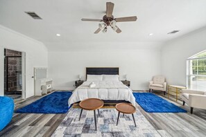 Un dormitorio con cama, dos mesitas de noche, ventilador de techo, alfombra azul y ventana con persianas.