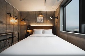 Een moderne hotelkamer met een groot bed, een houten hoofdbord, uitzicht over de stad, een wandlamp en een klein tafeltje met een stoel.