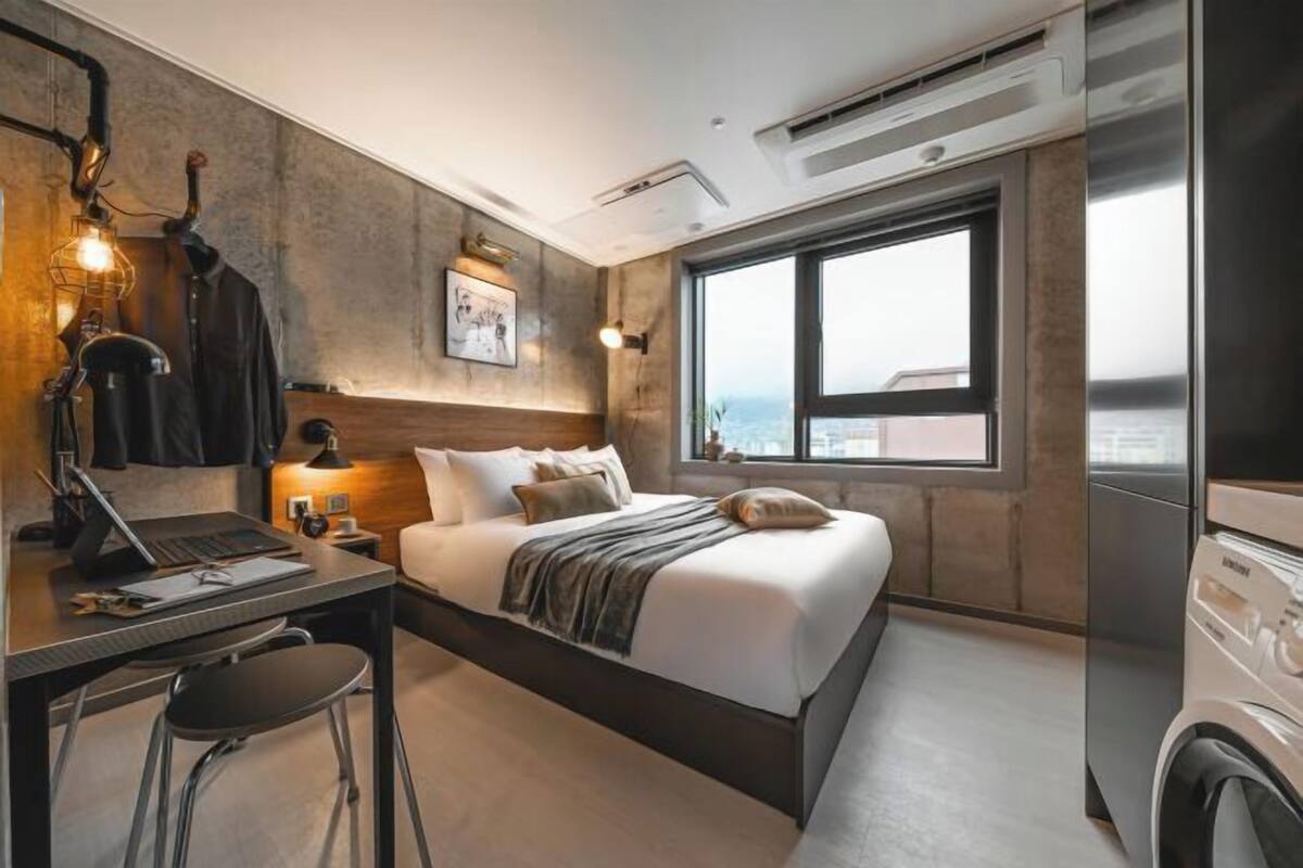 Une chambre d’hôtel moderne équipée d’un lit, d’un bureau et d’une chaise, d’une grande fenêtre donnant sur la ville et d’un lave-linge.