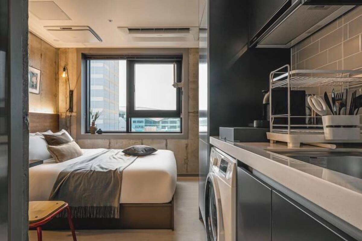 Une petite chambre comprenant un lit, un lave-linge et une fenêtre donnant sur des immeubles.
