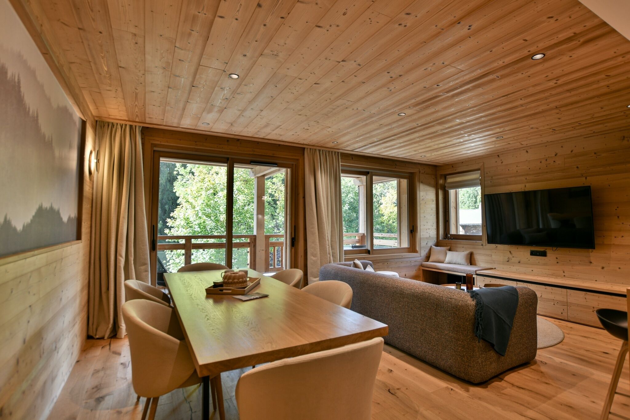 A cozy cabin interior with a wooden ceiling, large windows, a dining area, a sofa, and a TV.