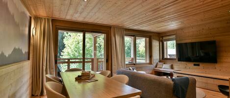 A cozy cabin interior with a wooden ceiling, large windows, a dining area, a sofa, and a TV.