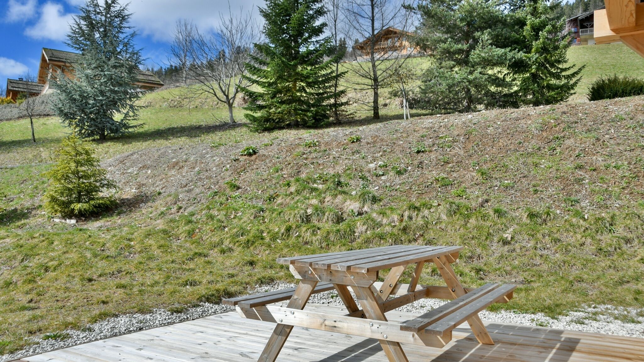 Une table de pique-nique en bois sur une terrasse en bois, avec une colline herbeuse et des arbres en arrière-plan.