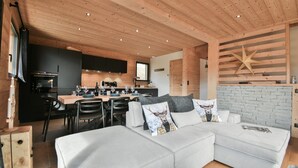Een moderne woonkamer met een hoekbank, een eethoek met een houten tafel en een keuken met donkere kastjes en een raam.