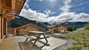 Een houten hutje met een terras en picknicktafel, omgeven door bergen en een heldere lucht.