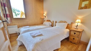 A cozy bedroom with two beds, wooden walls, a window with a view, and a nightstand with a lamp. - Refuge les Echelles E4 (Le Grand Bornand)