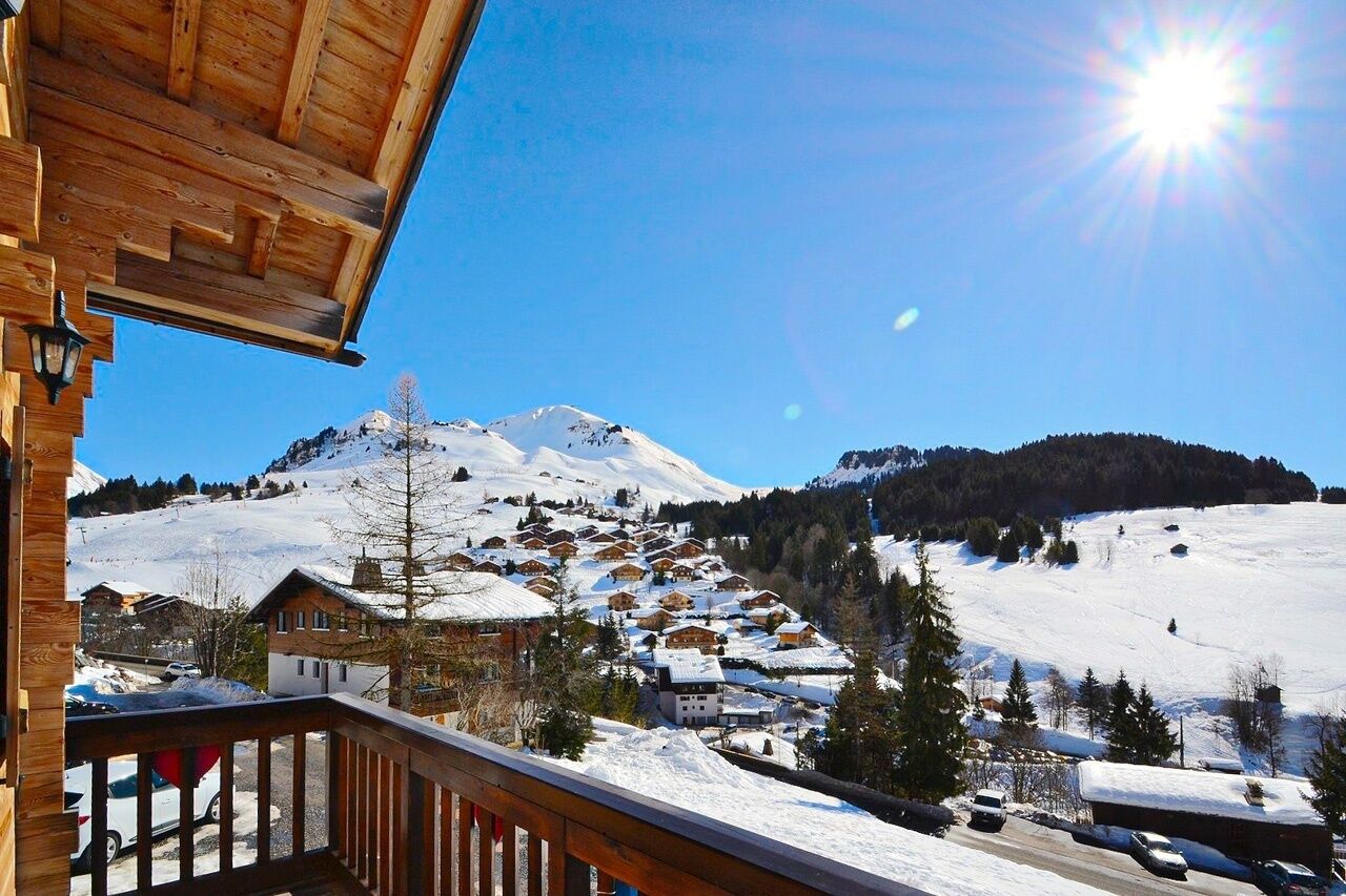 Un balcon en bois surplombant un village de montagne enneigé, avec ses maisons traditionnelles et un ciel d’un bleu limpide.