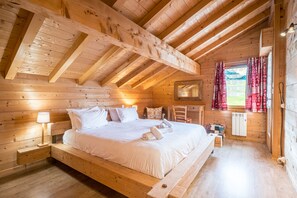 A wooden cabin bedroom with a loft bed, a bedside table with a lamp, a chair, and a window with curtains.