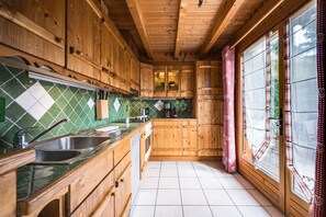 A wooden kitchen with a green and white tiled backsplash, wooden cabinets, and a window with checkered curtains.