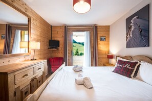 Habitación de hotel con paredes de madera, una cama con sábanas blancas, una silla roja, un televisor y un cuadro de un paisaje montañoso.