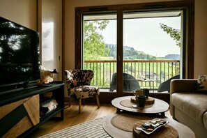 Un salon équipé d’un téléviseur, d’un canapé, d’un fauteuil, d’une table basse et d’un balcon offrant une vue sur la campagne.