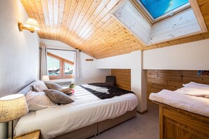 A bedroom with a wooden ceiling, a skylight, a bed with white bedding, a bedside table with a lamp, and a view of the outside through a window. - Florimontagne - Duplex 14 (Le Grand Bornand)