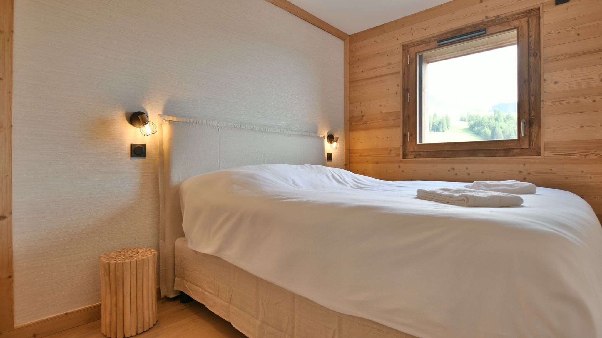 A neatly made bed with white linens and towels, two wall-mounted light fixtures, and a window with a view of greenery.