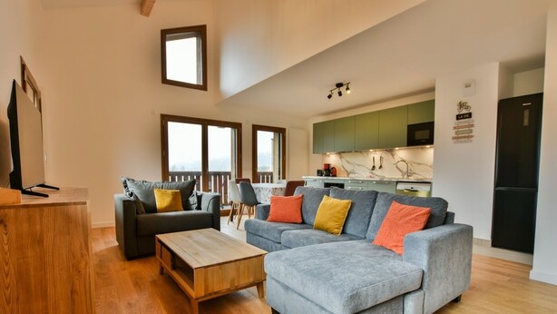 A modern living room with a grey sectional sofa, a wooden coffee table, and a TV on a wooden stand. There is a balcony with a view, a kitchen with green cabinets, and a black refrigerator. - Le Sève C201 - Three Bedroom Apartment, Sleeps 6 (Samoëns)