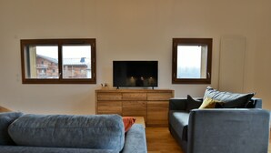 A living room with a TV on a wooden cabinet, two sofas, and two windows with views of buildings. - Le Sève C201 - Three Bedroom Apartment, Sleeps 6 (Samoëns)