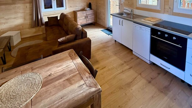 A cozy wooden cabin interior with a dining area, sofa, kitchen, and large windows. - Le Chalet de la Vernie | Private Jacuzzi | Hautes-Alpes (Saint-Léger-les-Mélèzes)