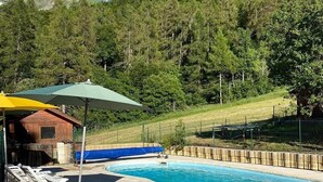 Une piscine entourée de chaises longues et de parasols, elle-même bordée d’une terrasse en bois et d’une colline boisée.