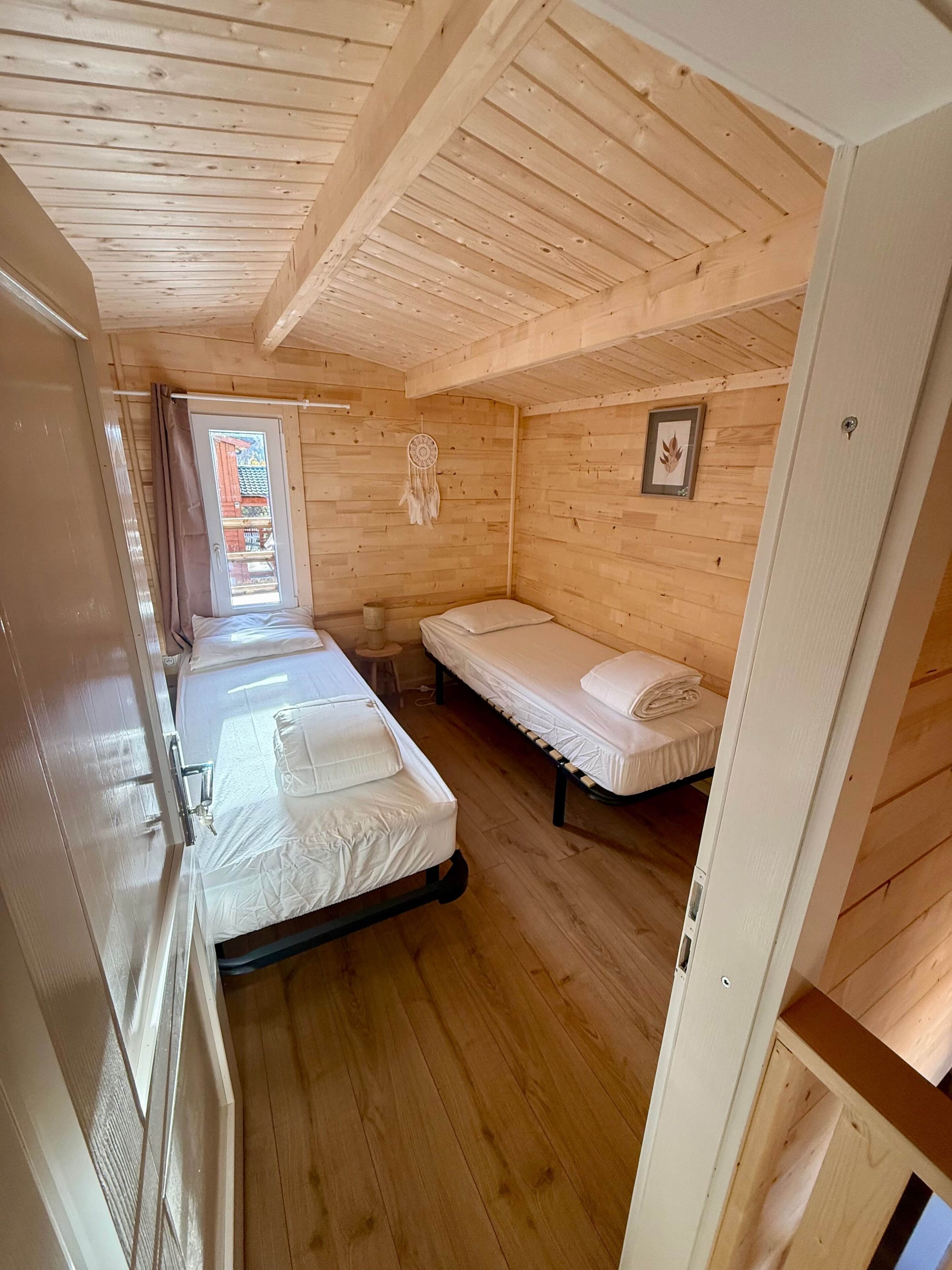 Een houten kamer met twee bedden, een raam met gordijnen, een oogsterminator en een ingelijst plaatje aan de muur.