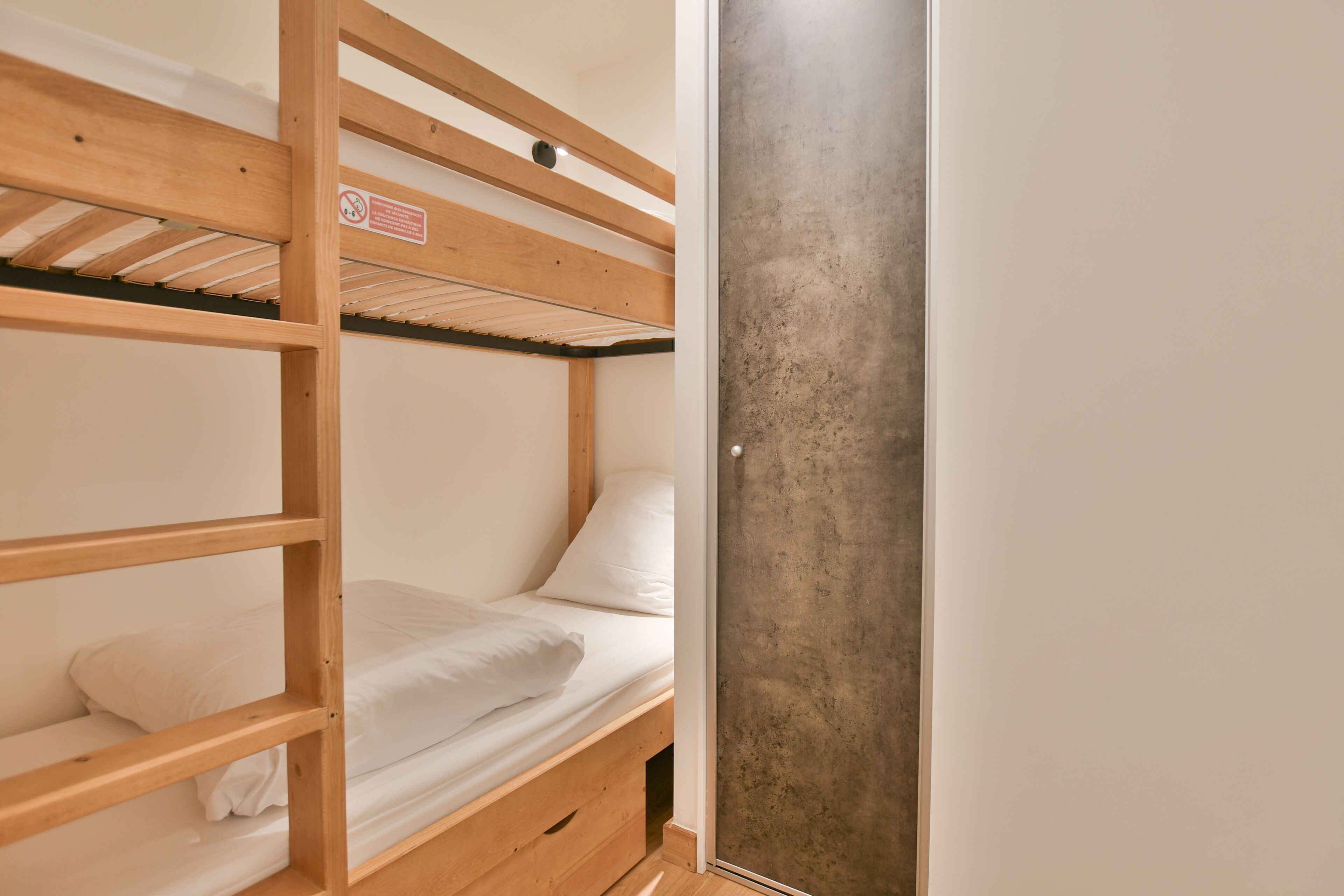 A bunk bed room with a wooden frame, white bedding, and a concrete wall.