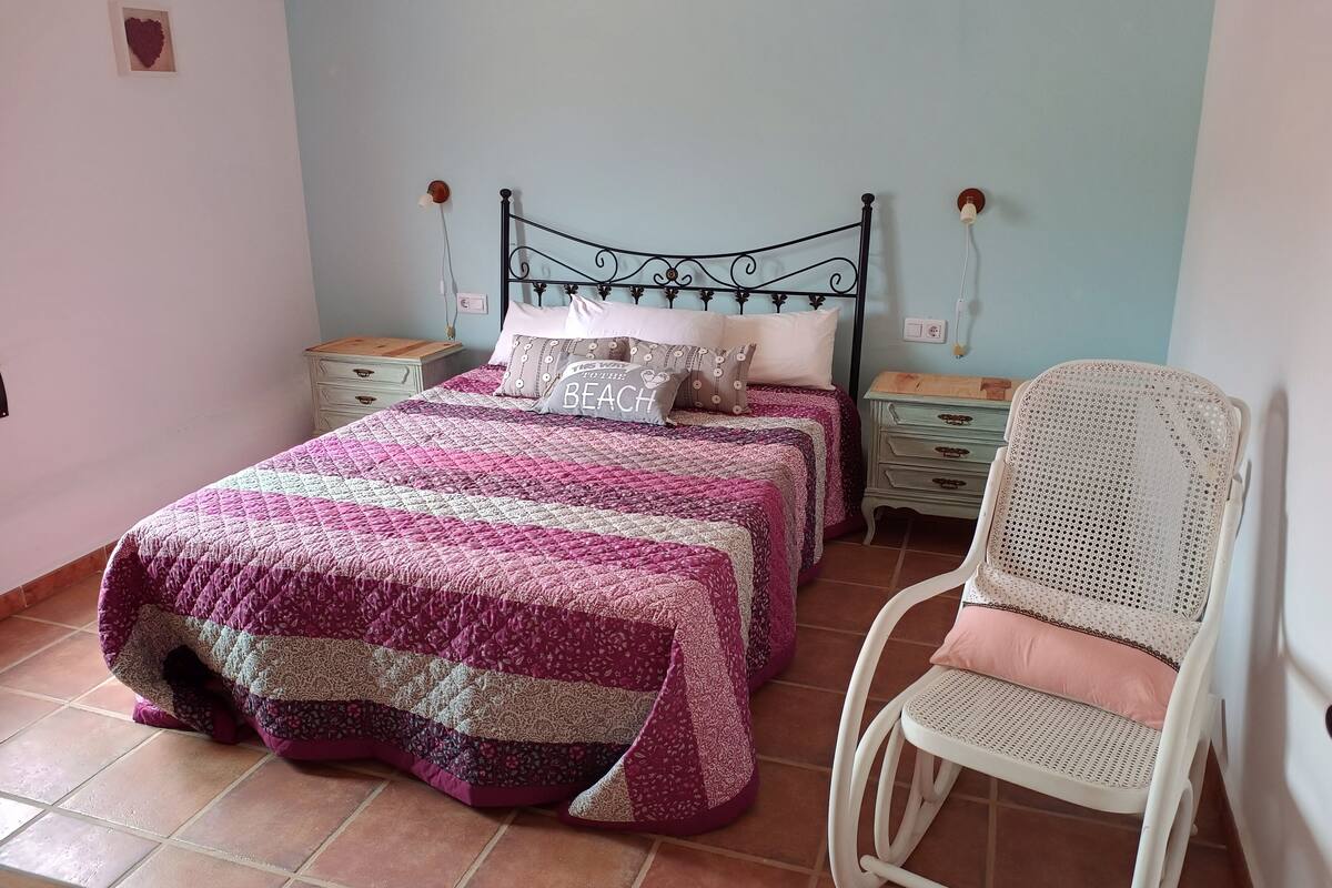 A bedroom with two beds, a white chair, and a nightstand with a lamp.