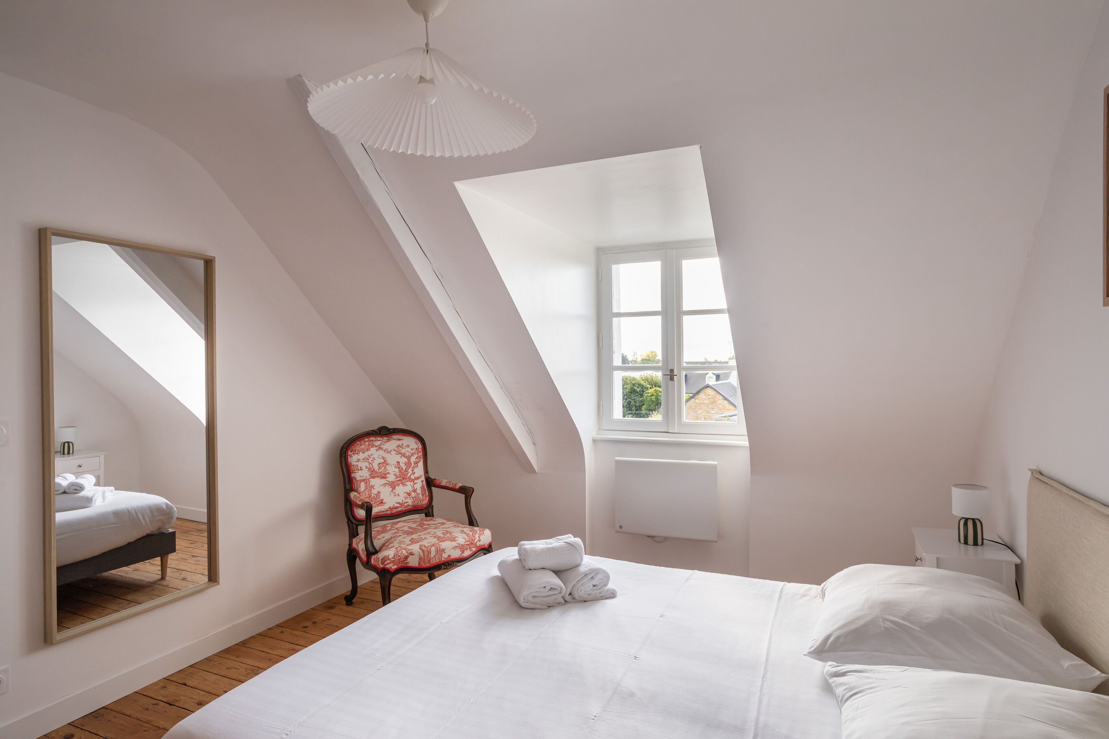 A bedroom with a large bed, a chair, a mirror, a window, and a ceiling light.