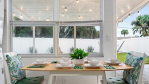 Un comedor con una mesa de madera dispuesta para cuatro personas, sillas blancas con cojines verdes y una planta en maceta sobre la mesa.