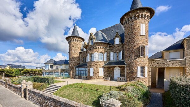 A castle-like building with multiple towers, a well-maintained lawn, and a stone wall.
