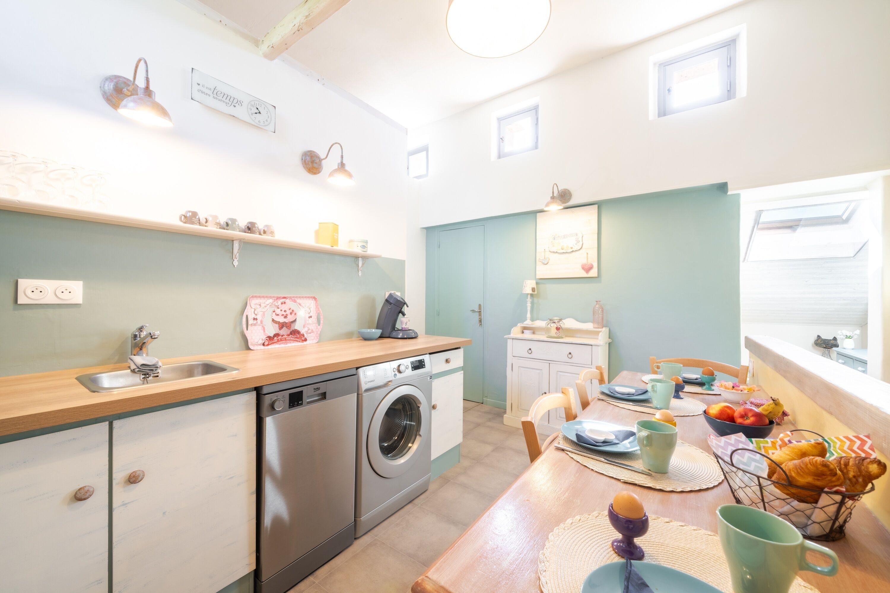 Une cuisine lumineuse équipée d’un lave-linge, d’un évier et d’un plan de travail en bois. On y trouve des chaises et une table avec des aliments pour le petit-déjeuner.