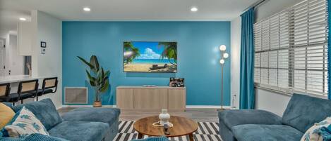 Una sala de estar moderna con una pared azul, un televisor de pantalla plana, un mueble de madera y una zona de comedor con sillas.