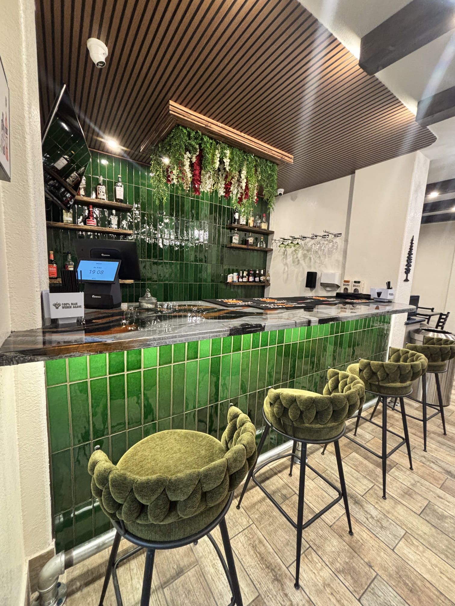 Un espace bar avec un mur recouvert de carreaux verts, une caisse enregistreuse et des tabourets de bar.