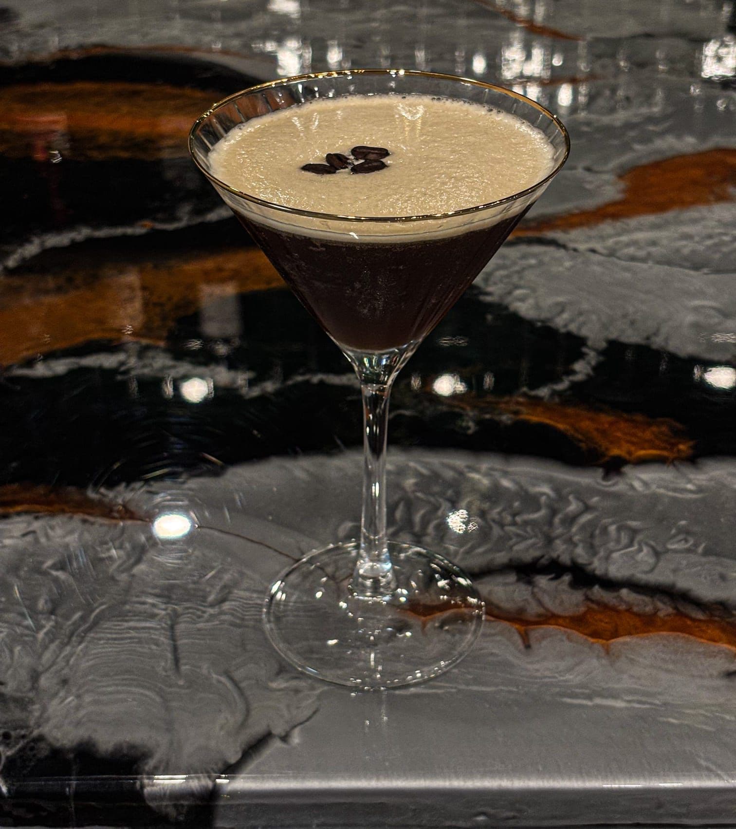 A martini glass with a dark liquid and a light-colored foam on top, placed on a reflective surface.