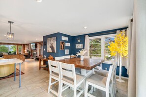 A dining area with a wooden table and white chairs, a kitchen in the background, and a view of trees through large windows. - Perfect Estes Park getaway-The Happy Place Estes,4BD,2Bath,HotTub,PetsOK,EV (Estes Park)