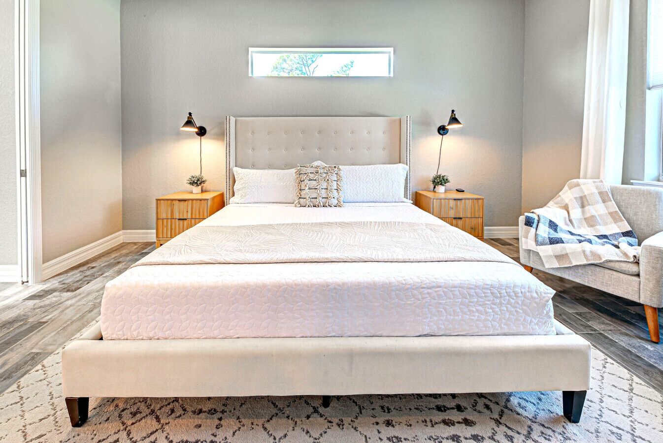 Een groot bed met een gestoffeerd hoofdeinde, twee nachtkastjes met lampen, een stoel met een geruit plaid en een ingelijst kunstwerk aan de muur.
